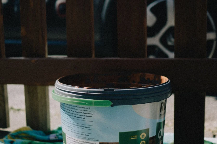 A paint bucket sits on a vibrant towel under sunlight beside a wooden fence.