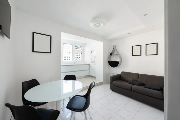 Interior of modern light house with glass table near chairs and couch with pillows near paintings and shelves on wall near entrance to kitchen with co