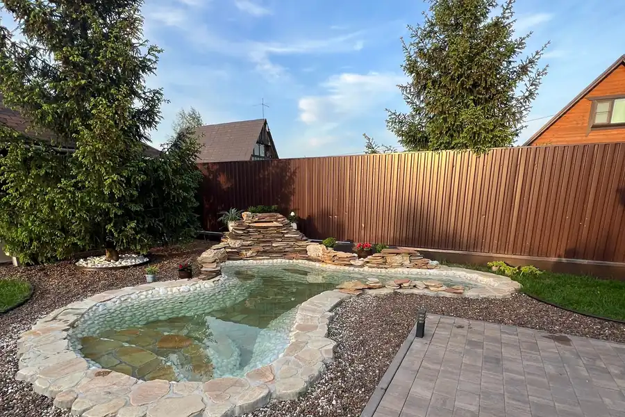 Serene backyard with stone pond, trees, and patio under a blue sky.